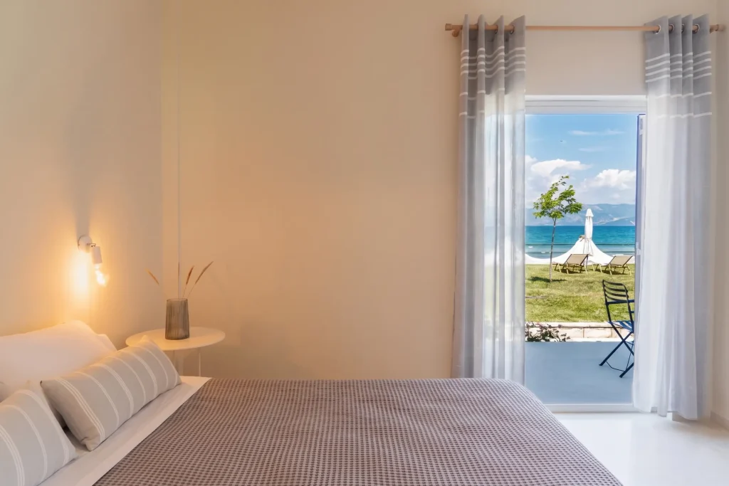 Ground-floor beachfront studio at Kamini Apartments Kavos with a double bed, warm bedside lighting, and direct walk-out access to a private terrace and the beach.