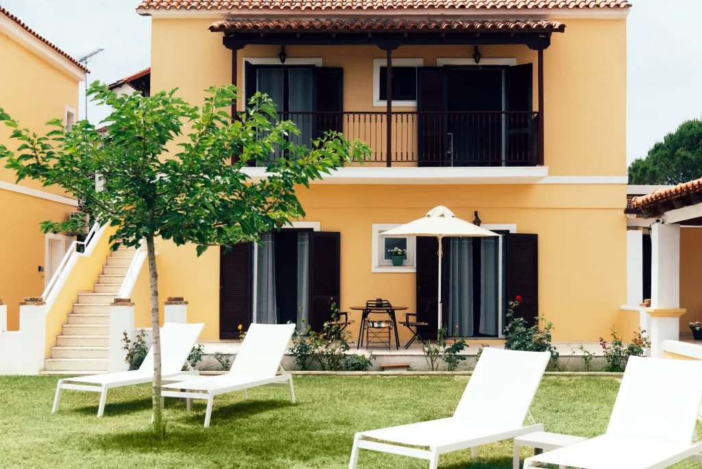 Exterior of Kamini Apartments Kavos featuring a traditional ochre building with private balconies, ground-floor terraces, and a green lawn with modern white sun loungers.
