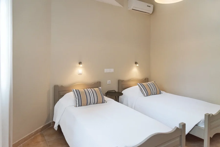Twin guest room at Kamini Apartments Kavos featuring two single beds with striped pillows, individual reading lamps, and wall-mounted air conditioning for guest comfort.