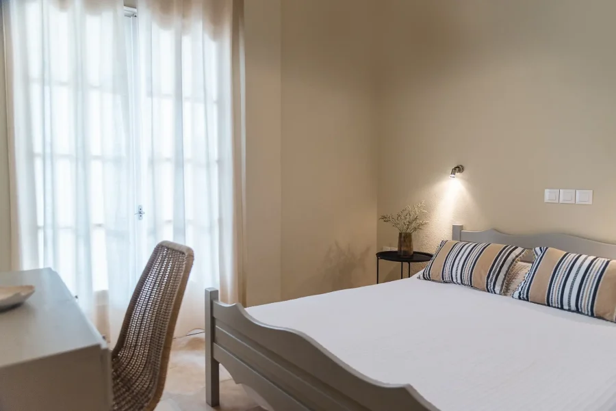 Bright double bedroom at Kamini Apartments Kavos featuring soft light from terrace doors, a comfortable bed with striped cushions, and a stylish wicker desk chair.