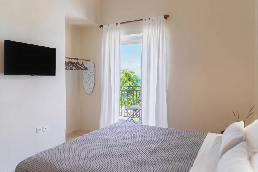 Bright sea-view studio at Kamini Apartments Kavos featuring a double bed, flat-screen TV, open clothing rack, and private balcony with garden views.