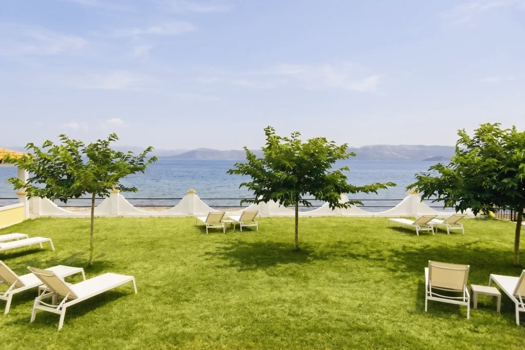 Relaxation area at Kamini Apartments in Kavos, Corfu, featuring white sunbeds on a lush green lawn just steps away from the Ionian Sea.