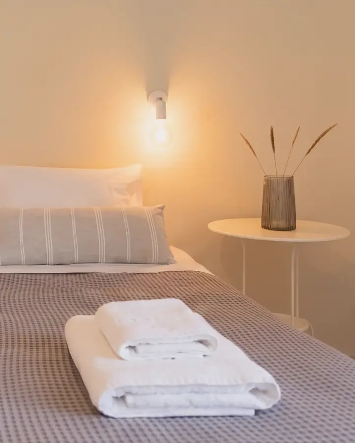 Freshly laundered white towels neatly folded on a made bed with crisp linens and warm bedside lighting at Kamini Apartments, Kavos.