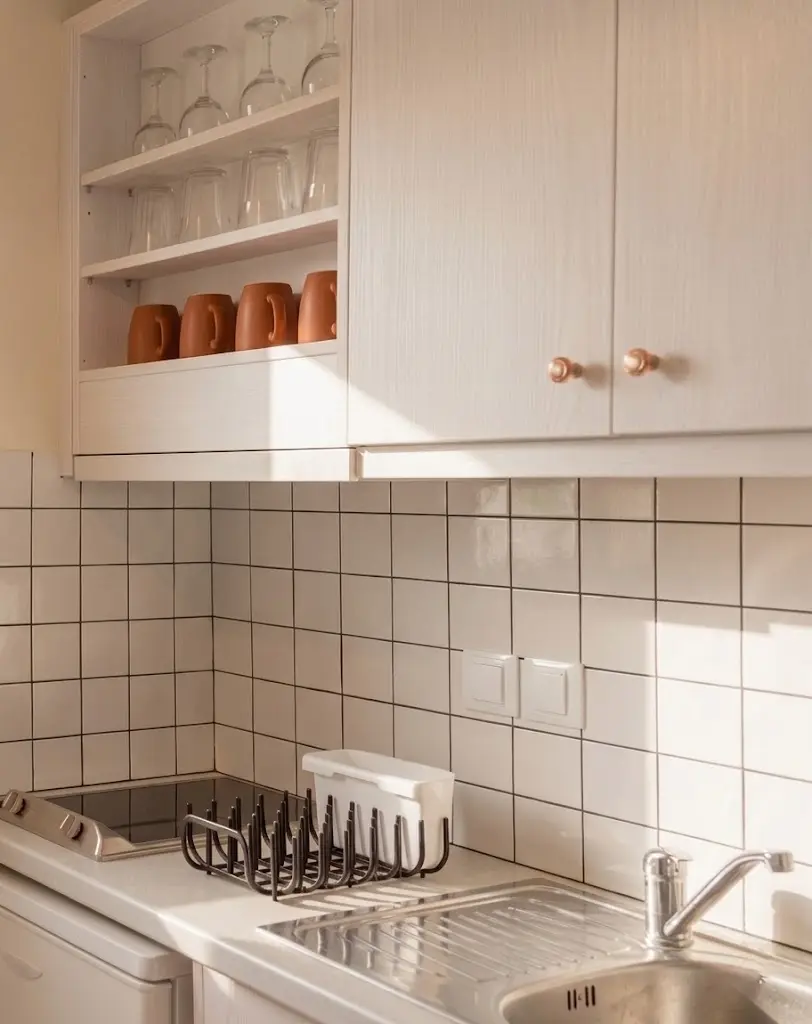 A clean, minimalist kitchenette at Kamini Apartments in Kavos, Corfu, featuring white tiling, organized glassware, clay mugs, and a stainless steel sink for self-catering.